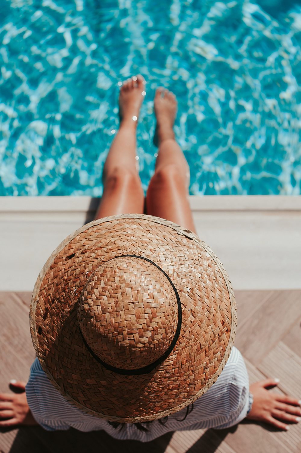A woman in a sunhat sitting by the edge of a pool.